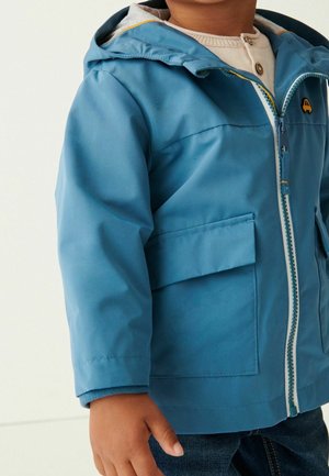 Enfant portant une veste de pluie à capuche bleue avec des poches avant et une fermeture éclair, sur un pull en tricot beige et un jean sombre.