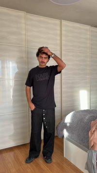 Mann i svart Karl Lagerfeld T-skjorte og løse bukser står innendørs med den ene hånden på hodet og den andre i lommen nær en seng med et grått teppe.