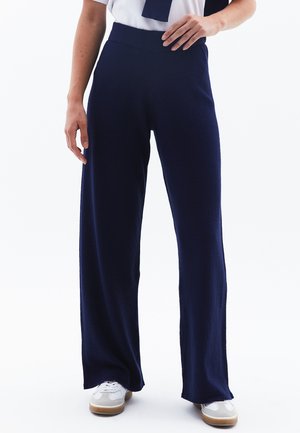 Pantalons larges bleu marine en tissu doux et texturé avec une taille haute, associés à des baskets blanches, renforçant le confort décontracté.