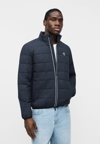 Άνδρας που φοράει ναυτικό μπουφάν με καπιτονέ μοτίβο και όρθιο γιακά, καθώς και ανοιχτόχρωμα μπλε τζιν, μπροστά σε απλό λευκό φόντο.