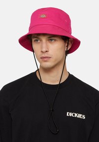Roze kape s plitkim obodom napravljene od čvrstog materijala, s remenom za bradu i šivenim detaljima. Model nosi crnu majicu s "DICKIES" logotipom.