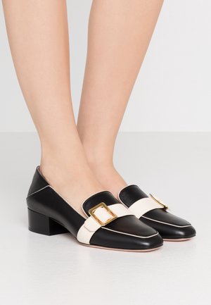 Schwarze flache Leder-Loafer mit weißen Riemen und goldenen Schnallen, getragen auf nackten Füßen vor einem schlichten Hintergrund.