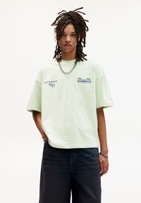 Lys grønt oversized t-shirt lavet af bomuld, med kontrasterende grafiske designs på forsiden. Båret med mørkeblå denim jeans.
