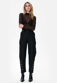 Transparante zwarte top met lange mouwen en gouden stippen, hoge hals. Zwarte op maat gemaakte broek met gouden knoopaccenten op de tailleband en enkels. Zwarte puntige laarzen.