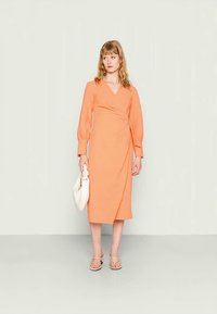 Vestido envelope em laranja vivo, com mangas bufantes longas e uma bainha até ao joelho. Combinado com uma bolsa branca e sandálias. Textura suave.