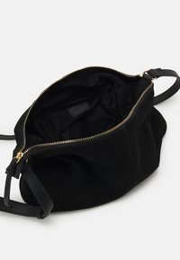 Sac à main en suède noir avec une fermeture éclair dorée, présentant un intérieur en tissu doux et une bandoulière en cuir noir ajustable. Étiquette intérieure visible.
