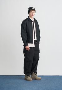 Μαύρο μπουφάν πάνω από ένα ανοιχτό ροζ πουκάμισο, σε συνδυασμό με μαύρο παντελόνι cargo και γκρι μπότες. Τα αξεσουάρ περιλαμβάνουν μαύρο σκουφί και γραβάτα.