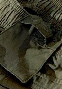 Zelená maskáčová tkanina s textúrovaným povrchom, obsahujúca vrecko na suchý zips a gumičkový pás. Detailný pohľad zdôrazňuje detaily.