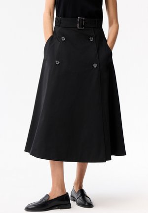Kobieta ubrana w czarną spódnicę midi o kroju litery A z guzikami z przodu i paskiem, w połączeniu z czarnymi skórzanymi mokasynami, stoi na tle białego tła.
