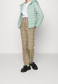 Light green quilted jacket, white t-shirt, plaid brown and green pants, black shoes with chunky sole, standing on a plain background.