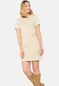 Beiges Kurzarmkleid mit Rundhalsausschnitt, ausgestattet mit zwei vorderen Taschen und Knopfakzenten. Sanfter Stoff und Oberschenkel-lange Länge.