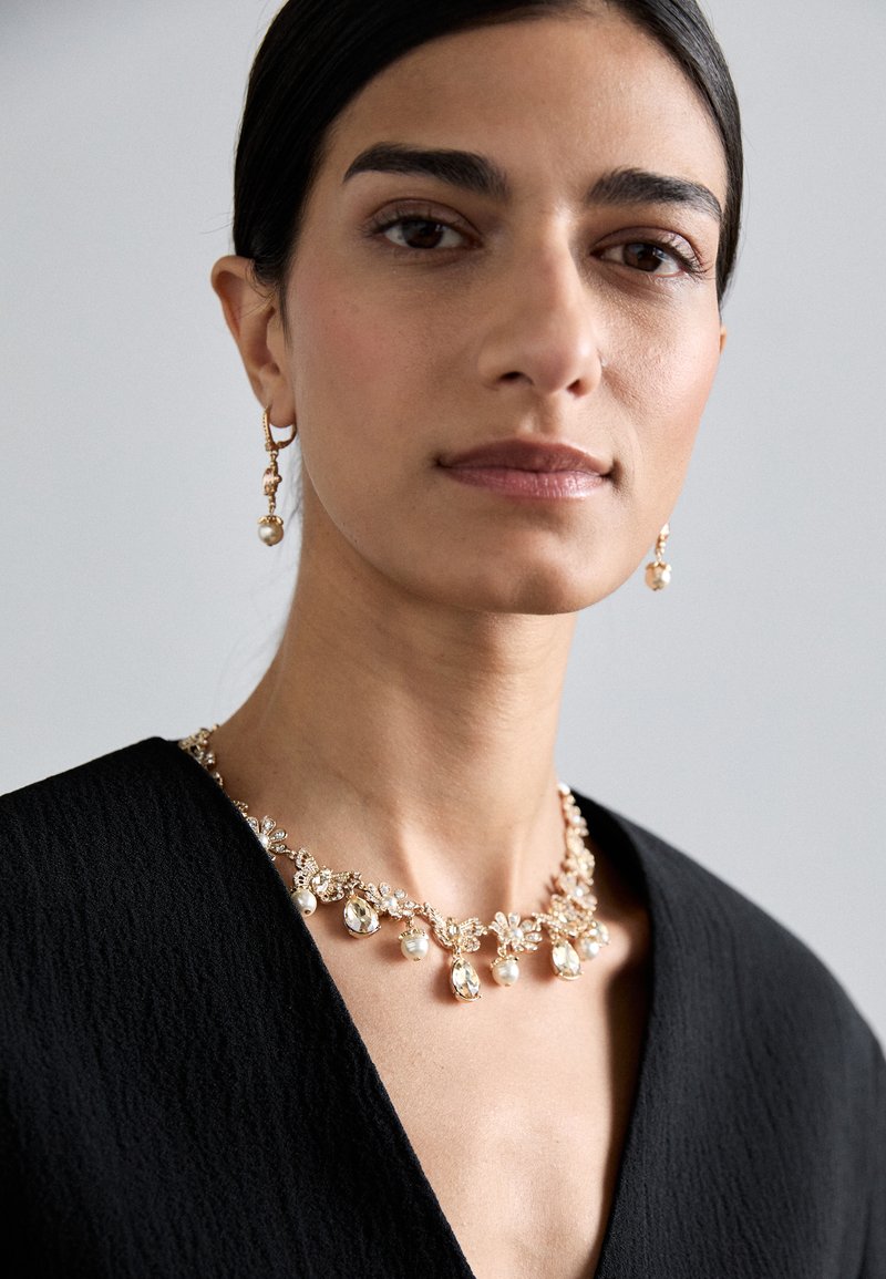 Halskette in Goldfarbe mit floralen Motiven, klaren Steinen und perligen Akzenten. Getragen mit passenden Ohrringen im Tropfenstil. Sichtbares schwarzes strukturiertes Oberteil.