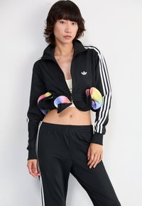 Veste de sport noire avec des détails de poignet multicolores, fermeture zippée à l'avant et bandes blanches sur les manches. Associée à un pantalon noir assorti.
