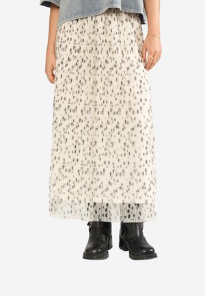 Femme portant une jupe longue plissée blanche avec des taches abstraites noires, des bottines noires et une veste courte en jean grise.