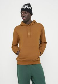 Καφέ φούτερ με κουκούλα, μπροστινή τσέπη τύπου καγκουρό και κορδόνι ρύθμισης. Συνδυάζεται με πράσινο κάτω μέρος. Το μοντέλο φορά επίσης μαύρο σκουφί με λευκές λεπτομέρειες.