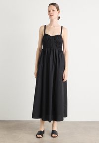 Robe maxi noire avec un corps ajusté, des bretelles réglables, des détails froncés à la taille et une jupe fluide. Texture lisse, design minimal.