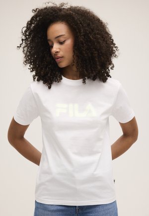 Frau mit lockigem Haar, die ein weißes FILA-T-Shirt und blaue Jeans trägt und mit den Händen hinter dem Rücken nach unten schaut.