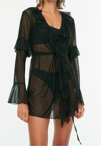 Black sheer cover-up with ruffled details, long sleeves, and a tie waist. Flared sleeve hems and a soft, lightweight texture.