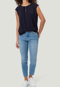 Blusa senza maniche blu navy con dettaglio a chiave sul davanti, abbinata a jeans aderenti blu chiaro e sneakers bianche. Design semplice e moderno.