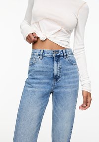 Bílý žebrovaný dlouhý rukáv s uzlíkovým předním detailem, spojený se světle modrými džíny s vysokým pasem a klasickým pětikapsovým designem.