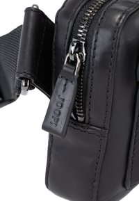 Zwarte leren tas met een gladde textuur, voorzien van zilverkleurige ritshardware en een merklabel aan de rits. Compact rechthoekig model met gestikte accenten.