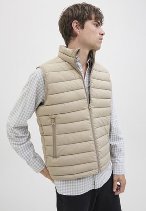 Hombre con chaleco acolchado beige sobre una camisa a cuadros azul claro, de pie con la mano en la cadera, mirando hacia su derecha contra un fondo blanco liso.