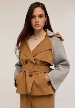 Junge Frau mit langen braunen Haaren trägt eine sandfarbene Jacke mit Gürtel, die graue Sweatshirtärmel hat, und passend dazu sandfarbene Hosen. Sie hat die Hände in den Taschen und einen neutralen Hintergrund.