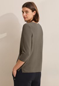 Olivgrön tröja med trekvartsärmar, strukturerad tyg, rund halsringning och sidofickor. Modellen står med händerna i fickorna.