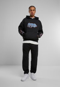 Sudadera negra con gráfico colorido en la parte delantera y las mangas, combinada con pantalones de chándal negros y zapatillas blancas. Tejido suave, ajuste casual.