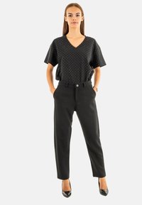 Haut noir à pois avec un col en V et manches courtes associé à un pantalon noir ajusté. Modèle portant des talons hauts noirs. Design simple et structuré.