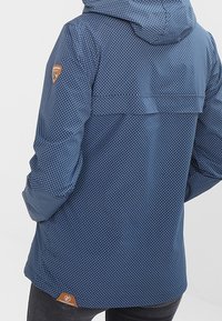 Granatowa kurtka w białe kropki, z kapturem i zamykaną na zamek kieszenią. Posiada fakturowany materiał oraz brązową łatę z logo na dolnej części.