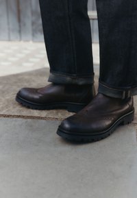Braune Leder-Stiefel mit rundem Zehenbereich, Brogue-Details und dicken schwarzen Gummisohlen. Dunkle Jeans mit umgeschlagenen Bündchen.