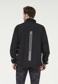 Veste de sport noire avec un col montant, détail réfléchissant "ENDURANCE" au dos et accents texturés sur les manches.