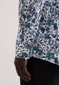 Camicia bianca con un motivo floreale in blu scuro e teal, con polsino abbottonato e manica arrotolata. Tessuto di stoffa liscio.
