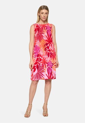 Frau steht und trägt ein ärmelloses knielanges Kleid mit roten, rosa und orangefarbenen tropischen Blattmustern sowie goldene Riemchensandaletten vor weißem Hintergrund.