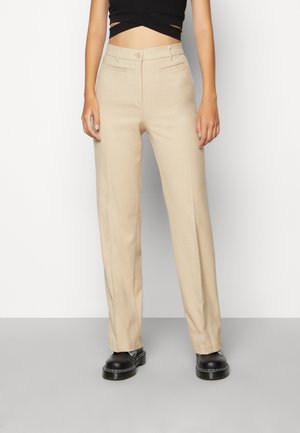 Persoon die beige rechte broek, zwart cropped top en zwarte stevige veterboots draagt, staand tegen een eenvoudige grijs-witte achtergrond.