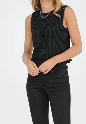 Mujer con un top negro sin mangas con rayas y botones, y vaqueros negros, con un collar plateado, manos juntas delante.