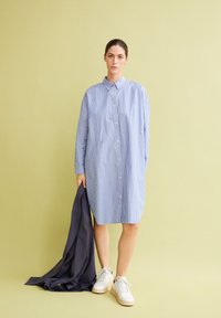 Robes chemises rayées bleues et blanches, avec boutons, col, manches longues. Associée à une veste sombre tenue d'une main et des baskets blanches.