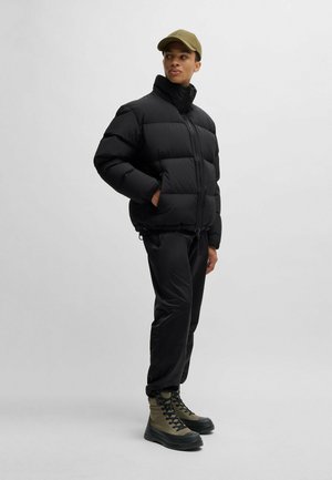 Schwarze Steppjacke mit hohem Kragen, Reißverschluss und seitlichen Taschen. Kombiniert mit schwarzen Hosen und grünen Schnürstiefeln. Neutraler Hintergrund.