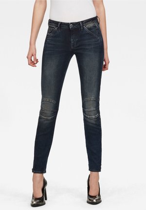 Jeans de mezclilla azul oscuro con un corte ajustado, que presentan acentos en los paneles de las rodillas y un sutil desgastado. Combinados con tacones altos negros brillantes.