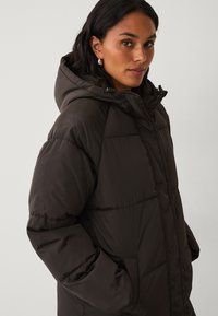 Bruine pufferjas met een capuchon, voorzien van een doorgestikt ontwerp, een volledige rits en zijzakken. Gladde textuur en oversized pasvorm.