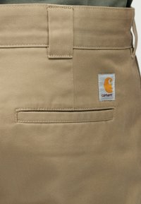 Μπεζ παντελόνι εργασίας με ευθεία γραμμή, που διαθέτει πίσω τσέπη και ραμμένη ετικέτα με το πορτοκαλί και λευκό λογότυπο Carhartt. Λείο, ανθεκτικό ύφασμα.