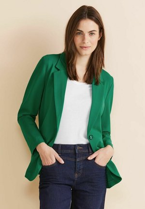 Groene blazer met een gladde textuur, sluiting met één knop en twee voorzakken, gecombineerd met een witte top en donkere spijkerbroek.