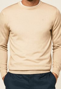 Pull beige en maille avec un col rond et des manches longues, présentant une texture lisse et des poignets ajustés.