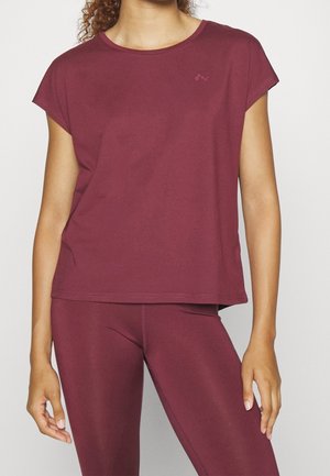 Femme portant un haut ample à manches courtes de couleur bordeaux et des leggings ajustés assortis, debout devant un fond clair uni.