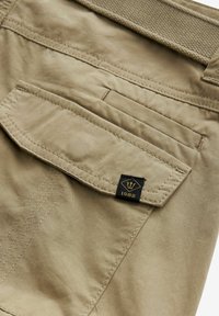 Khaki spodnie z tylną kieszenią z klapką oraz małą czarną metką z logo korony i rokiem 1982. Gładka tekstura.