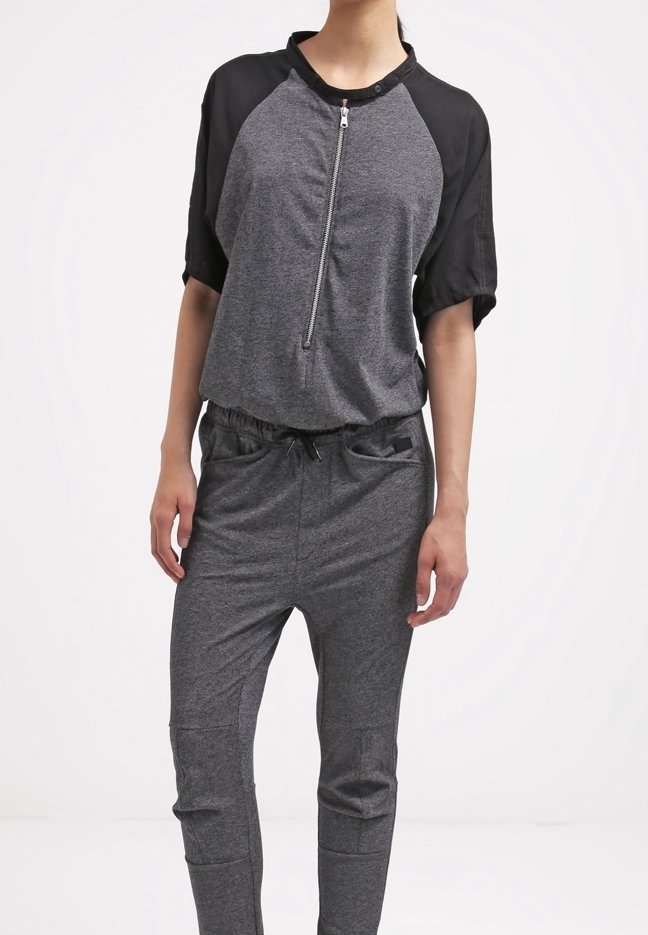 Femme portant un haut zippé manches courtes gris et noir avec un pantalon gris à cordon assorti, debout devant un fond blanc.