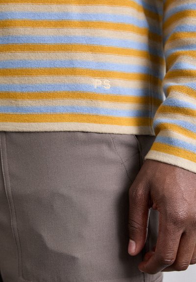 Sárga, kék és bézs csíkos pulóver; kötött textúra; az alján "PS" logó található; szürke szövet nadrággal párosítva.