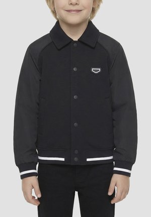 Dijete u crnoj jakni na kopčanje s bijelim prugastim manšetama i rubom, stoji ispred jednostavne svijetle pozadine.