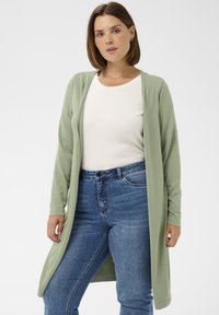 Sieviete, kas ģērbta gaiši zaļā garajā kardigānā, baltā T-kreklā un zilās džinsās, stāv uz vienkārša baltā fona.